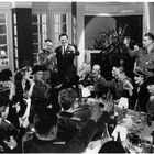Menschen in Uniformen und Abendkleidung feiern und stoßen bei einem großen Tisch an, fröhliche Stimmung, zahlreiche Gläser.