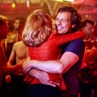 Ein Mann in einem blauen Shirt umarmt glücklich eine Frau in rotem Glitzerkleid inmitten einer feiernden Menschenmenge.