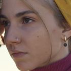 Ich kann keine Texte im Bild sehen. Eine Person mit Nasenring und Ohrring blickt zur Seite, trägt ein gelbes Tuch.