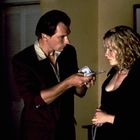 Ein Mann reicht einer Frau Geldscheine. Beide stehen in einem Raum. Eine Skulptur und ein Bild sind im Hintergrund sichtbar.