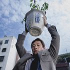 Ein Mann hebt einen schweren Blumentopf über seinen Kopf. Im Hintergrund sind Wohngebäude und Wolken am Himmel.