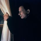 Eine Person in einem dunklen Raum zieht einen Vorhang beiseite und schaut hinaus.