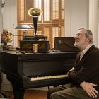 Ein Mann sitzt vor einem Mason & Hamlin Klavier, darauf ein Edison Phonograph. Er wirkt nachdenklich, im Raum herrscht Vintage-Stil.