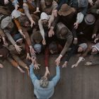 Viele Menschen in alten, ärmlichen Kleidern strecken die Arme aus in Richtung einer Person auf einem Holzsteg.