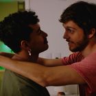 Zwei Männer stehen nah beieinander in einem Raum, der im Hintergrund grün beleuchtet ist. Einer trägt einen Stern-Ohrring.