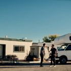 Zwei Männer vor einem weißen Wohnmobil mit "indie campers"-Schriftzug, daneben ein Truck und ein Gebäude mit Getränkeautomat.