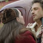 Keine sichtbaren Texte. Eine Frau hält einen Mann mit einer Pistole am Kragen eines alten Autos fest.