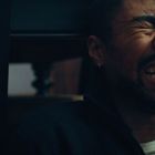 Kein Text. Ein Mann sitzt am Boden, lacht oder weint, mit geschlossenen Augen und offenen Lippen.