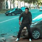 Ein Mann mit blutigen Händen steht vor einem verbeulten Auto auf einer Straße, umgeben von weiteren Autos.