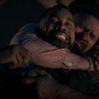 Zwei Männer ringen miteinander. Der vordere Mann hält eine Granate, der hintere umklammert ihn von hinten und wirkt angespannt.