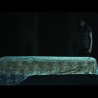 Kein Text erkennbar. Eine Frau steht neben einem Tisch, der mit einem Tuch bedeckt ist; im Hintergrund dunkle Umgebung, zwei Kerzen.