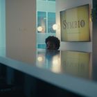 Text: "SYMBIO". Ein schmaler Raum mit einem langen, glänzenden Tisch, Stühlen, einem Fenster und einer Küchenzeile mit Pflanzen.
