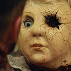 Eine zerbrochene Puppe mit blonden Locken und einem zerschlagenen Auge, umgeben von Rissen und Schrammen im Gesicht.