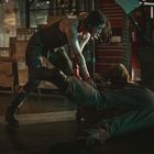 Ein blutüberströmter Mensch kämpft mit einer anderen Person in einem dunklen Lagerhaus voller Kartons und Werkzeugen.