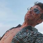 Ein Mann mit blutverschmiertem Gesicht und offenem Hemd schaut nach oben, vor einem hellblauen Himmel.