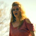 Ich kann keine Texte erkennen. Eine Frau in einem roten, schulterfreien Oberteil schaut erschrocken. Im Hintergrund sind unscharfe Bäume.