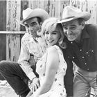 Zwei Männer in Cowboyhüten und eine Frau in einem gemusterten Kleid lachen vor einer Holzwand.