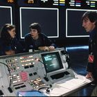 Konsole mit Bildschirmen, Tasten und Zahlen, zwei junge Personen und ein älterer Mann sprechen miteinander in einem Kontrollraum.