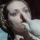 Eine Person mit blauen Augen wird von einer behandschuhten Hand auf die Nase gedrückt. Die Haare sind kunstvoll gesteckt.