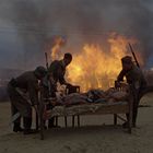 Drei Soldaten tragen ein Bett mit einer Person darauf vor einem großen Feuer. Der Himmel ist dunkel und rauchig.