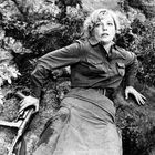 Keine sichtbaren Texte. Eine Person liegt in Uniform an einem felsigen Hang, hält ein Gewehr in der Hand.