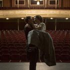 Ein Paar umarmt sich auf einer leeren Theaterbühne unter einem großen Kronleuchter.