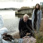 Polizei in Schutzanzügen am Ufer, ein Mann kniet neben einer Leiche, eine Frau steht daneben, Himmel bewölkt.