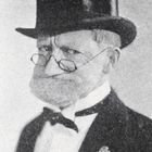 Keine sichtbaren Texte. Ein Mann trägt einen Zylinder, Brille, Fliege und Anzug mit Blumenansteckern.