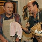 Zwei Männer, einer trägt eine Schürze und hält ein Handtuch, der andere hat einen Korb. Sie stehen in einem Raum mit Holztür.