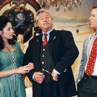 Ich kann den Text im Bild nicht erkennen. Drei Personen in eleganter Kleidung in einem Raum mit historischer Dekoration.