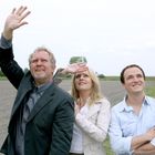 Drei Personen schauen in die Ferne: ein Mann hebt die Hand, eine Frau schützt ihre Augen, ein weiterer Mann lächelt.