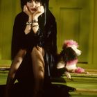 Frau in dunkler Kleidung auf Stufen, den Kopf in die Hände gestützt. Neben ihr sitzt ein Pudel mit rosa Details.