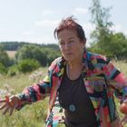 Keine Texte. Eine Frau in bunter Jacke hält einen Apfel in einer grünen Wiese, bewaldeter Hintergrund.