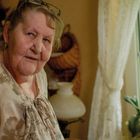 Eine ältere Frau sitzt in einem gemütlichen Raum mit Pflanzen am Fenster und einem Traumfänger. Sie trägt eine Brille und eine gemusterte Bluse.