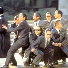 Sieben Männer in Anzügen, einige mit Sonnenbrillen, stehen in dynamischen Posen auf einer Treppe.