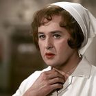 Eine Person in einem Krankenpfleger-Outfit mit weißem Kopftuch in einem Flur, schaut nachdenklich.