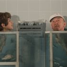 Zwei Köpfe schauen aus Aquarien hervor, in denen Fische schwimmen. Ein älterer Mann trägt eine Mütze, eine Frau lächelt.