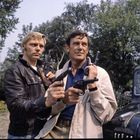 Zwei Männer stehen nebeneinander, beide mit Revolvern. Im Hintergrund sind Bäume und ein altes Auto zu sehen.