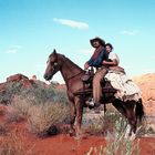 Zwei Personen reiten auf einem Pferd durch eine trockene, rote Wüstenlandschaft mit Büschen und sonnigem, klarem Himmel.