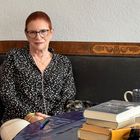 Eine Frau sitzt auf einem Sofa mit Kissen. Ein Stapel Bücher, ein Stift und eine Tasse stehen auf einem Tisch.