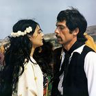 Ich sehe keinen Text. Ein Mann und eine Frau stehen sich gegenüber in traditioneller Kleidung, umgeben von Menschen und Natur.