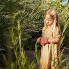 Keine Texte sichtbar. Ein blondes Mädchen in einem gelben Kleid steht ernst inmitten von hohem Gras und Sträuchern.