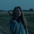 Eine Frau steht in einer dämmerigen Landschaft und hält ein Gewehr über ihrer Schulter.