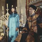 Drei Menschen in traditionellen, asiatisch inspirierten Gewändern in einem prunkvollen Raum mit vielen Kerzen.