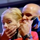 Ein Mann hält eine Frau fest und spricht in ein Telefon. Die Frau wirkt erschreckt, ihre Hand ist an ihrem Mund.