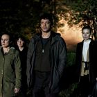 Vier Menschen stehen im Dunkeln im Freien, ein Mann und drei Frauen, mit ernstem Ausdruck, umgeben von Bäumen.