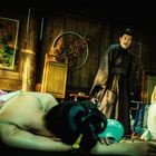 Ein Mann in traditioneller Kleidung steht, während zwei Menschen am Boden liegen, einer hat Schnittwunden auf dem Rücken.