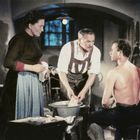 Ein Mann sitzt oben ohne auf einem Hocker, während zwei andere Personen bei ihm stehen. Sie befinden sich in einer Werkstatt.