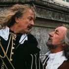 Zwei Männer in historischen Kostümen stehen sich gegenüber; einer hält einen Degen. Beide haben lange, graue Haare.