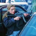 Eine Frau mit blonden Haaren hält ein Gewehr. Sie lehnt sich aus einem Autofenster in einer urbanen Umgebung.